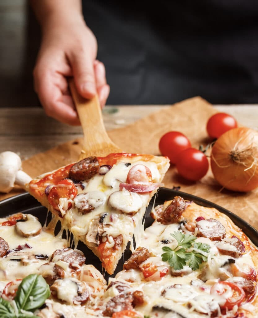 Pizza uppdelat i bitar nygräddad på bord. En person håller upp en bit med stekspade
